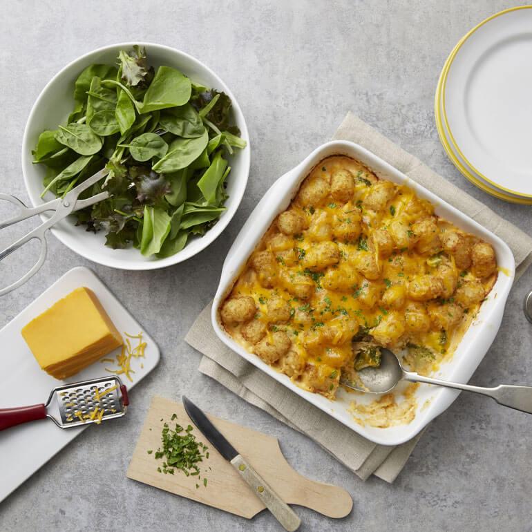 Recipe image of cheesy broccoli chicken potato casserole next to a bowl of salad, a block of cheese and a cutting board