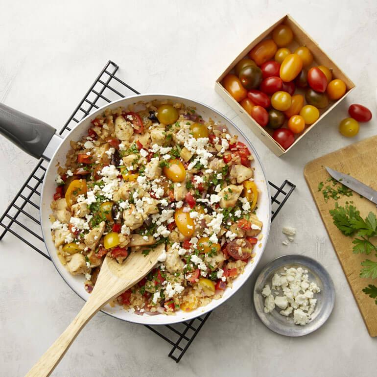 Skillet of Greek chicken and riced cauliflower next to a box of tomatoes, a cutting board and a bowl of feta for the recipe